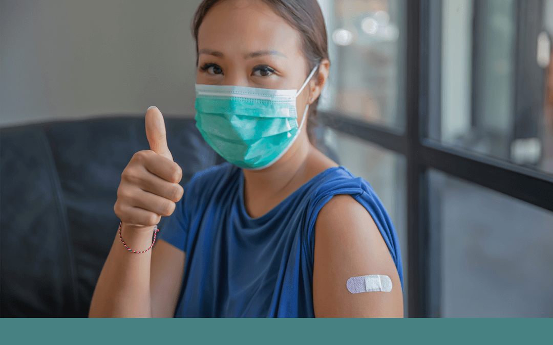 Photo of an asian woman wearing a mask with a bandage on her arm and holding her thumb up.